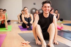 A man sits on a yoga mat and smiles as he participates in a yoga class as a part of his fitness therapy
