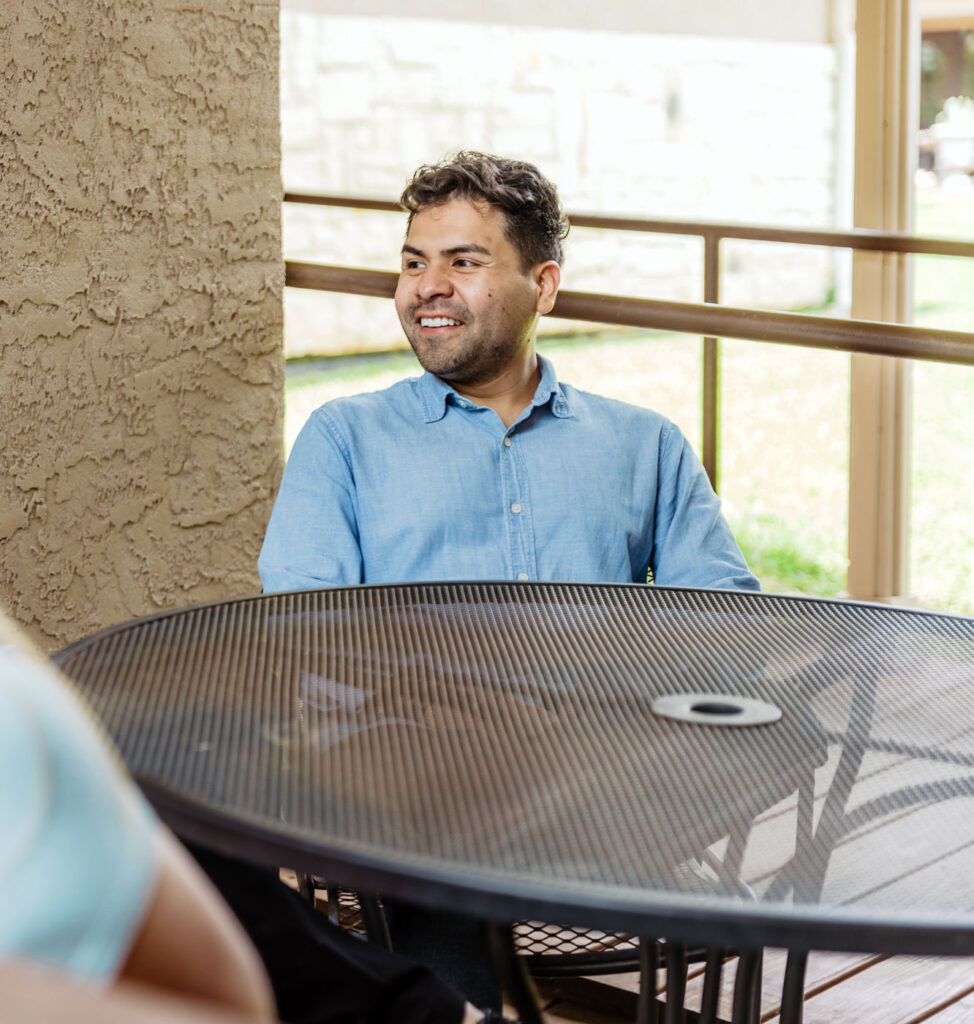 image of yong man seated at table with another person on a sunny balcony and smiling