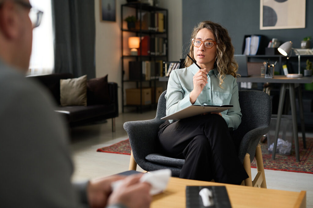 woman with glasses sits in a chair with note pad on her lap and talks to another person sitting across from her