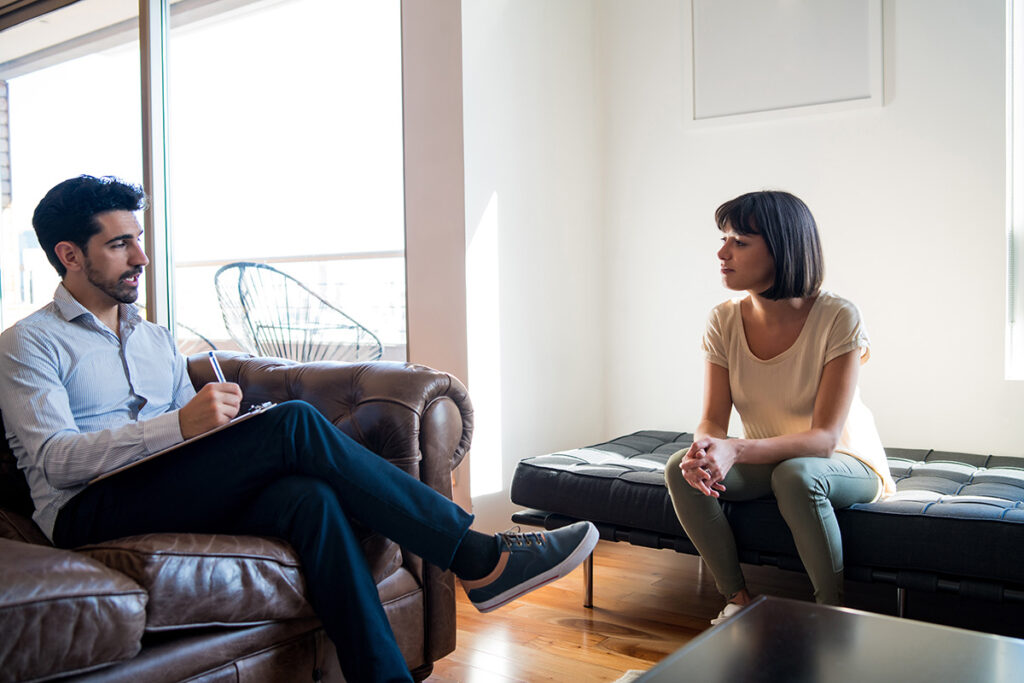 woman sits and looks at guy on a couch who is talking and writing on a notepad