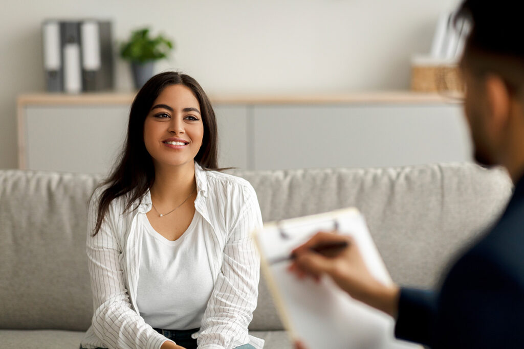 smiling woman with dark hair sits on couch while talking to therapist in her heroin addiction treatment program