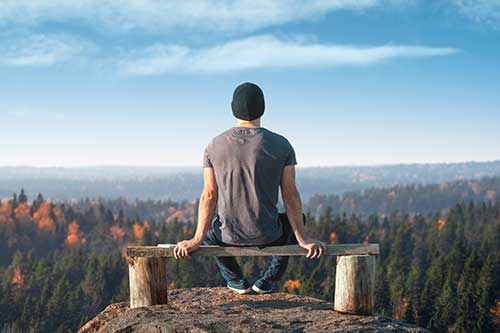 young man seated on bench looking out over beautiful wooded mountainous landscape and reflecting on shame resiliency