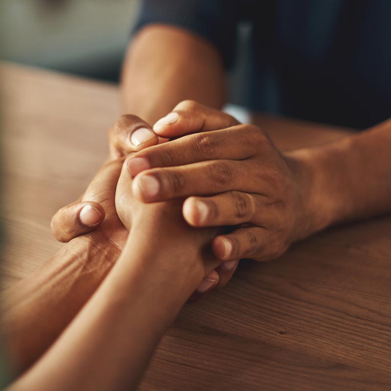 close up image of two people's hands grasped together on a table in a gesture of comfort.